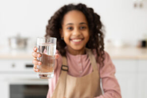 Closeup Shot Of Cute Little Black Girl Holding Glass Of Water In Hand And Giving It At Camera, Enjoying Drinking Healthy Refreshing Beverage, Sitting At Table In Kitchen At Home, Selective Focus